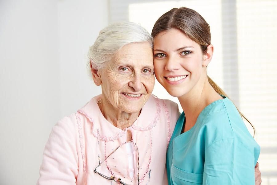 Elderly woman in a pink cardigan smiling beside her caregiver in teal scrubs